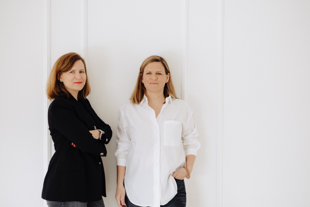 Zwei Frauen stehen in einem hellen Raum, eine trägt einen schwarzen Blazer und die andere ein weißes Hemd. Beide wirken professionell und selbstbewusst.